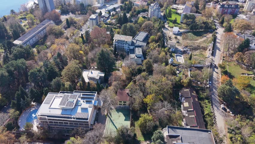 Aerial view of a small town surrounded by hills and autumn forest, showing residential buildings, roads, and natural landscape under clear skies.