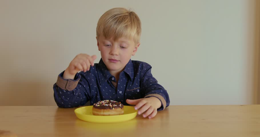 A blond boy dressed in a blue shirt has a donut with colorful sprinkles in front of him on a yellow plate. He is learning to count how many colorful pieces there are.