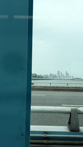 Scene captured from a vehicle in motion as it crosses a sprawling bridge spanning a river. In the far-off horizon, the urban skyline emerges, dotted with towering skyscrapers