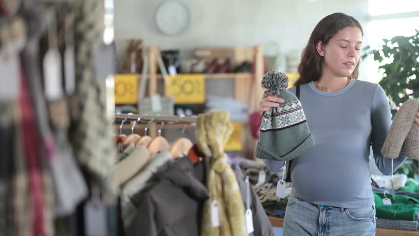 Selective young pregnant customer holding knitted hat for cold season in retail outlet