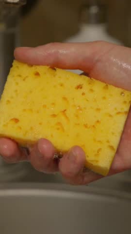 Vertical close-up of a hand squeezing a dish sponge filled with thick foam, with soap bubbles dripping down. The shot conveys cleaning routines, texture details and a tactile household moment. 4k