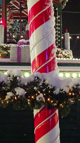 A giant candy cane stands proudly adorned with snow at a vibrant winter market. Twinkling lights and decorations create a cheerful atmosphere as night sets in