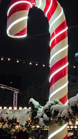 A giant candy cane stands proudly adorned with snow at a vibrant winter market. Twinkling lights and decorations create a cheerful atmosphere as night sets in.