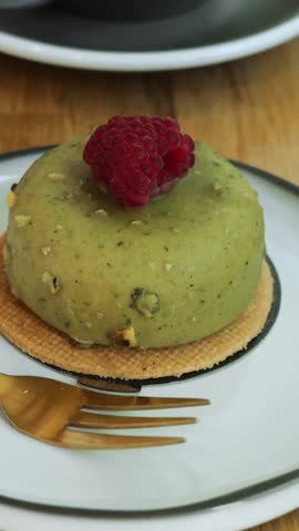 Green pistachio cake with fresh raspberry on top and cappuccino cup on wooden table. Healthy dessert and foamy coffee with cream heart served in coffee shop vertical video