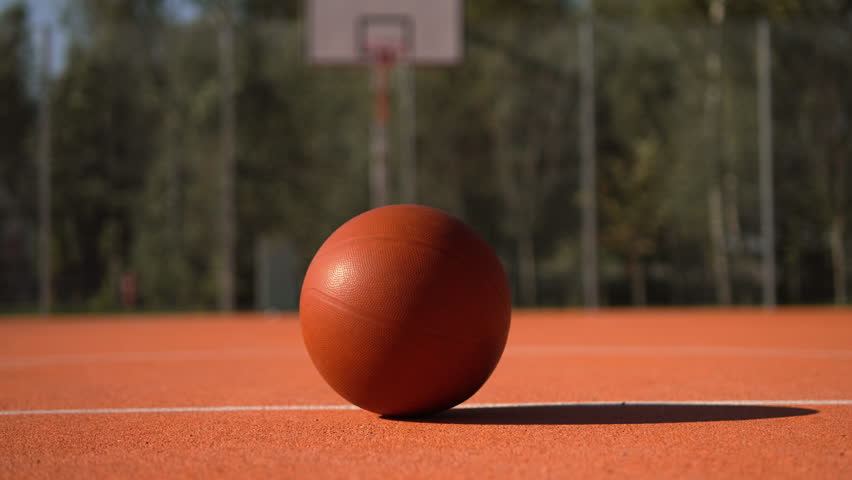 Basketball lies motionless in center of new rubberized outdoor basketball court, opposite backboard and hoop. Camera pans back away from orange ball on sunny day.