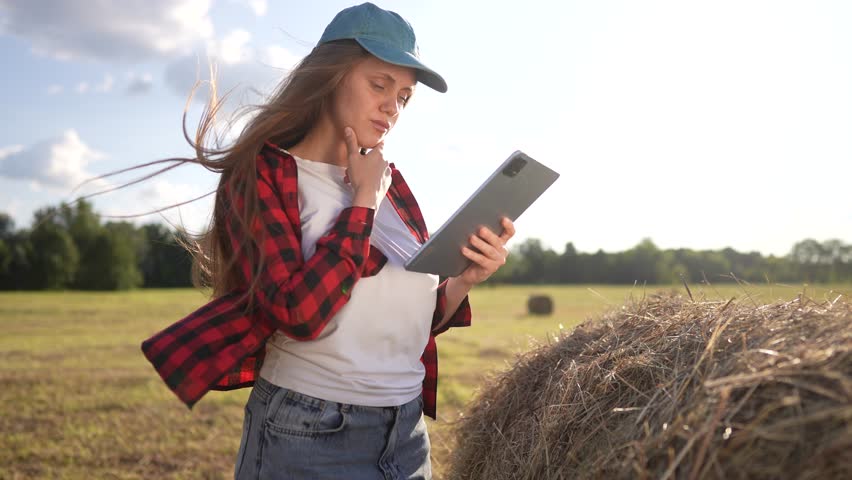 A woman farmer in a red plaid shirt stands next to a hay bale, using a tablet in the field. The woman interacts with the tablet, surrounded by hay bales and a plaid shirt.