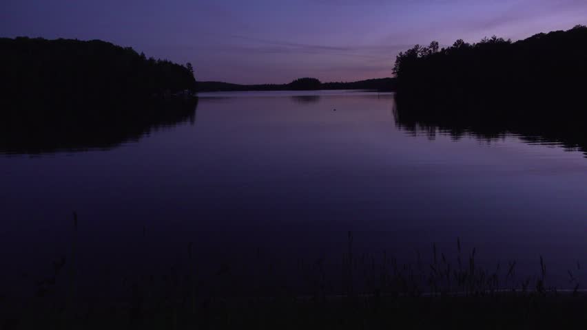 A calm lake reflecting a purple sunset and silhouetted trees in a rural area.