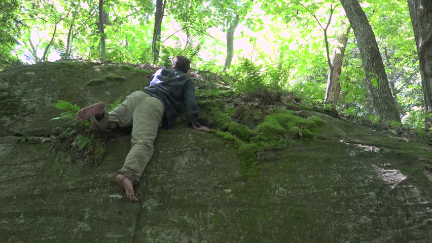 A low-angle view of a young barefooted man climbing a large boulder in a forest during the summer.