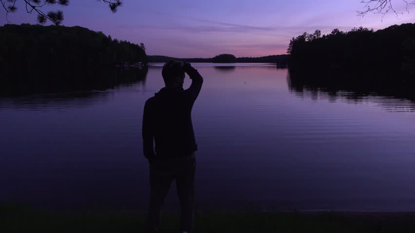 A silhouetted young man stands by a tranquil lake at dusk, watching the sunset over the water. Gentle ripples and reflected purple skies create a peaceful, contemplative mood.