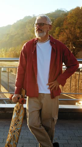 Retired man shows confidence with longboard on footbridge at sunrise. Active and healthy mature male with beard enjoys sportive lifestyle in modern city
