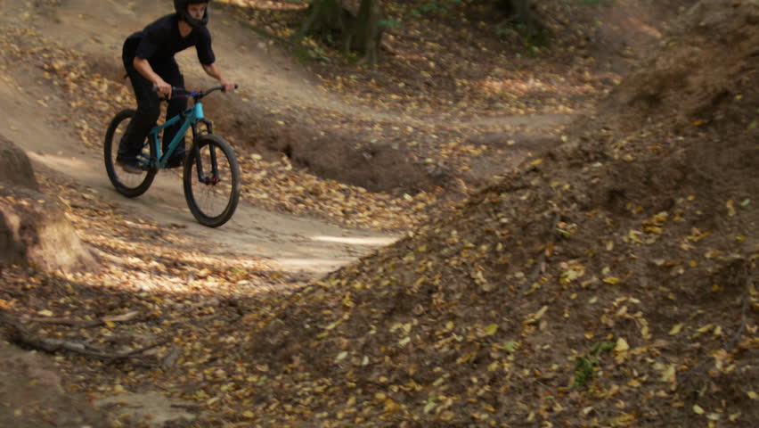 Skilled cyclist in helmet jumps with bicycle over hills ramp at training in extreme park slow motion. Young sportsman tricks with dirt bike in autumn forest