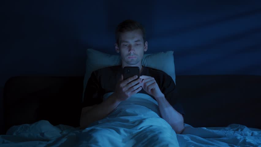 A tired man lies awake in a dark bedroom, staring at his glowing smartphone and doom scrolling late at night despite his growing fatigue. Digital addiction or insomnia concept. Copy space 