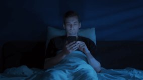 A tired man lies awake in a dark bedroom, staring at his glowing smartphone and doom scrolling late at night despite his growing fatigue. Digital addiction or insomnia concept. Copy space  - Powered by Shutterstock - Get 15% off with code: PIKWIZARD15