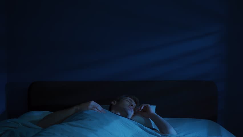 A shocked man abruptly awakes and sits up in bed at night, startled and breathing fast because nightmare disturb his already troubled sleep. Sleep disorder, insomnia and night terror. Space for text