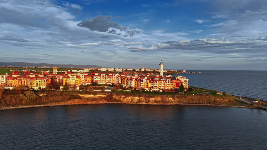 A stunning coastal landscape features a lighthouse standing tall beside colorful buildings, with waves gently lapping at the shore under a vibrant sky at sunset.