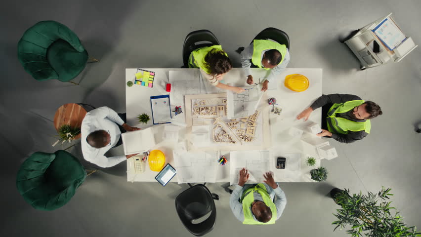 Top down view zoom out of property developers and architects in meeting reviewing blueprints and structural designs. Team discusses project approval and city planning for housing growth. Camera A.