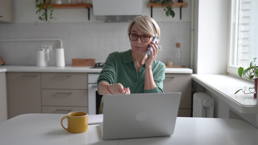 Middle aged businesswoman working multitasking on laptop, writing notes in notebook and gets distracted with smartphone, checking messages, answering call, having troubles with concentration and ADHD.