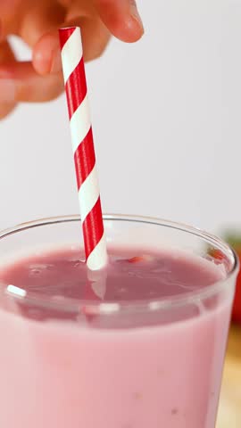 Hand stirs creamy pink fruit smoothie in glass using red striped straw, bright natural lighting