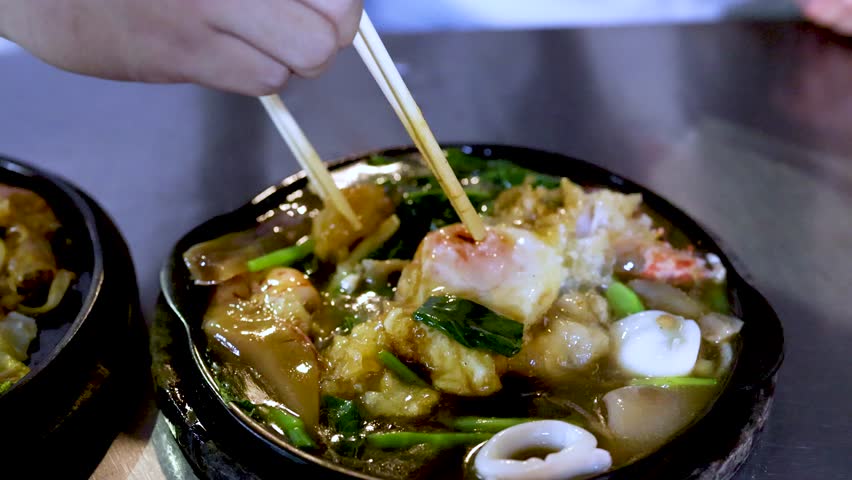 Chopsticks lift seafood pad thai noodles from a sizzling hot plate under bright lighting.