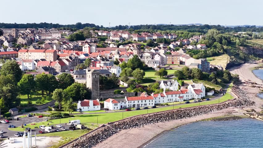 Drone glides above Fife coastal town, revealing beachfront houses, historic tower, and rugged shoreline