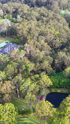 Drone glides above forested luxury estate, revealing houses, pool, pond, and lush greenery