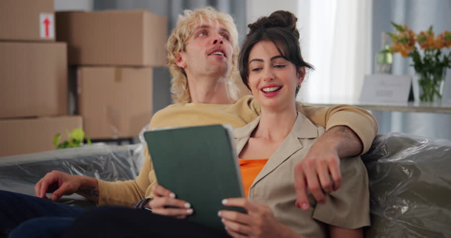 Happy, couple and planning with tablet in new home for online shopping, decor app and decision. Moving, talking and people with tech on sofa for interior design, furniture inspiration and remodeling