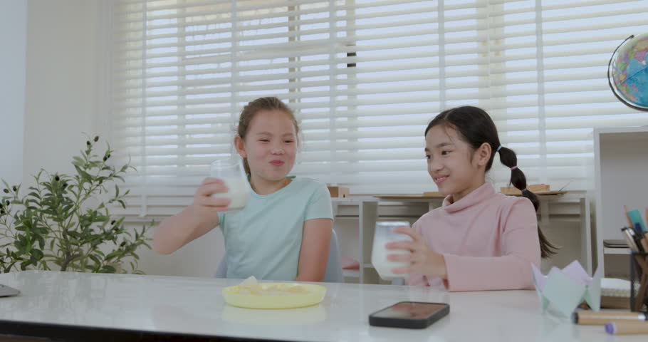 Two cute Asian sister girls clinking fresh milk glasses and drinking together cheerfully at a home table, representing good health and growth.