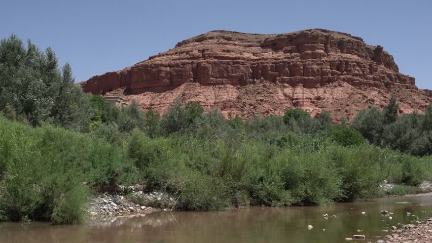 A desert mountain with no vegetation under a blue sky. In the foreground, there is a small river with lots of greenery on the other side.