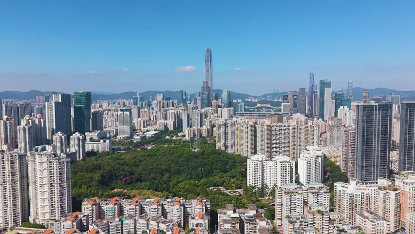 Aerial view of Shenzhen’s skyline featuring Ping An Finance Centre rising above dense urban districts. Modern high-rises, green park areas, and distant mountains under a clear blue sky. China, UHD.