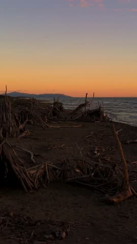 Driftwood tipi standing on the sandy beach shore in Tuscany under glowing evening skies. Maremma aerial