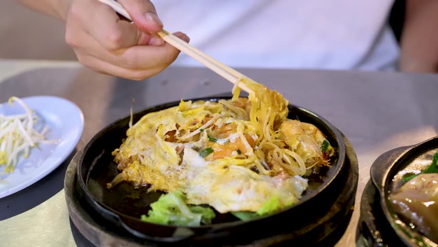 Hand lifts hot Pad Thai noodles with chopsticks from sizzling plate in bright indoor setting.