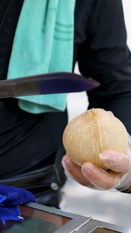 Gloved vendor skillfully slices coconut open with machete at outdoor market, natural daylight, close-up