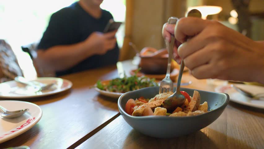 Person mixes spicy Thai noodle salad with utensils in bright, casual restaurant setting, shallow focus