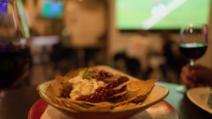 Hand reaches for nachos with salsa, sour cream, wine, sports game on TV, warm lighting.