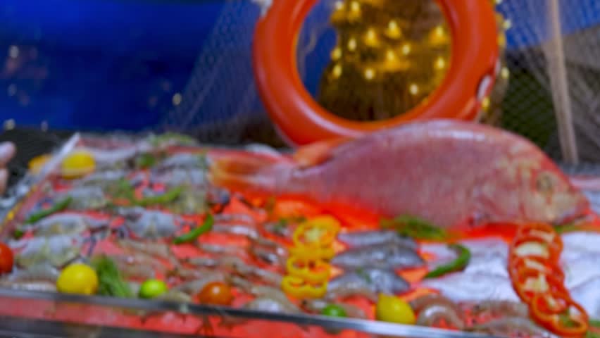 Hand refreshing a raw seafood buffet display featuring red snapper, blue crabs, and shrimp. Vibrant market setup with lemon garnishes, vegetable decoration, and nautical life buoy background.