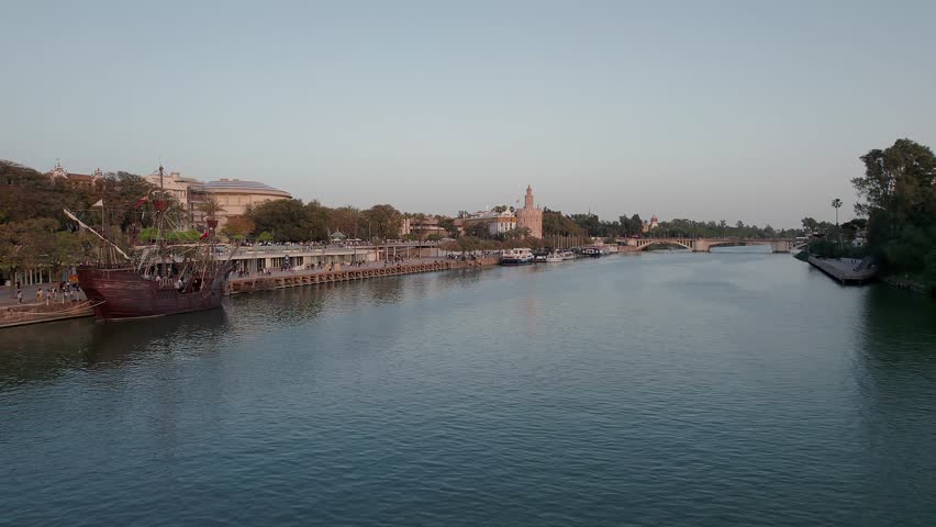 Guadalquivir River in Seville, capturing a replica Galleon docked alongside the lively promenade, with the historic Torre del Oro and the Puente de San Telmo defining background of Andalusian skyline