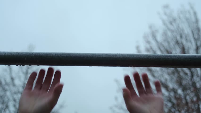 Fixed close-up of hands gripping a wet pull-up bar under a grey winter sky.