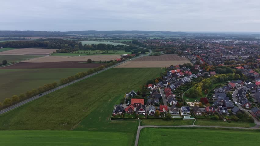 Aerial view of German village surrounded by open fields, winding residential streets and early autumn colors blending into the rural landscape. Wide shot. Hilly rural landscape in Germany.