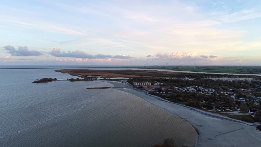 Aerial view of Dutch IJsselmeer coastline during ebb tide, showing shallow water patterns, wind turbines in distance. Calm shoreline under soft evening light in Netherlands. Wide shot.