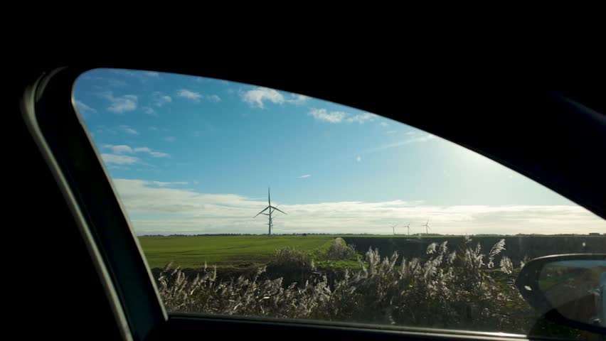 Static shot green fields with a wind turbine through a car window on a bright sunny day