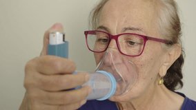Elderly woman with breathing problems using an inhaler with a spacer and an oxygen mask to take her medication, pressing the pump to release the spray into the chamber before inhaling - Powered by Shutterstock - Get 15% off with code: PIKWIZARD15
