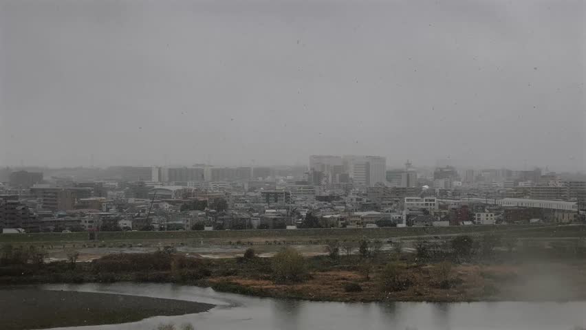 View of Futakotamagawa on a snowy day, overlooking the Tamagawa River and buildings across the water. Snowflakes drift through the grey, overcast sky, capturing a rare and serene winter scene.