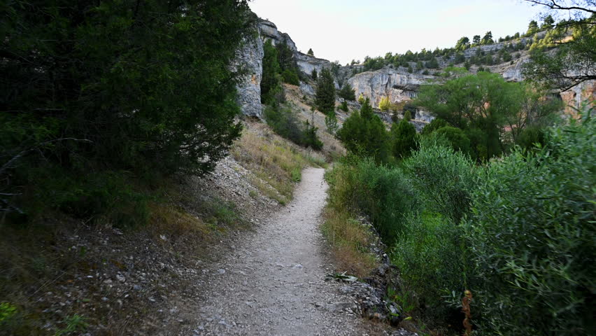 A first person view follows a narrow trail with a wooden railing in the canyon of Rio Lobos, leading toward the hermitage of Saint Bartolome through rocky cliffs and dense green vegetation. 
