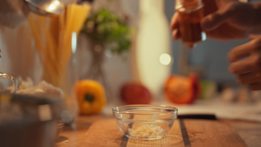 In a warm kitchen, someone adds seasoning from a small spice jar into a bowl with minced garlic to create a flavorful marinade. This action takes place during meal preparation