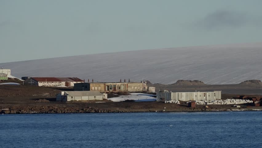 Remote polar coastal outpost with weathered buildings and shipping containers on a barren tundra shoreline beside calm icy waters, framed by a snow-dusted slope and pale sky