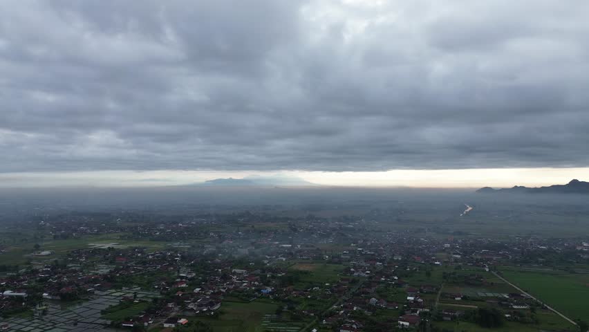 Aerial drone view of farmland and village under heavy clouds with distant mountains on the horizon. Calm and moody landscape.