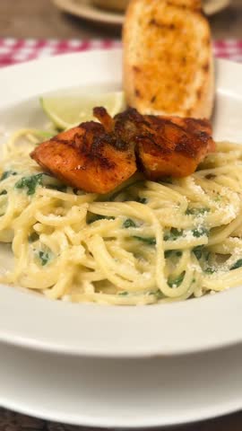 Close-up footage of creamy spinach pasta being mixed with a fork and spoon,topped with grilled salmon.Warm restaurant setting, Asian hand, appetizing food motion perfect for cooking or dining themes.