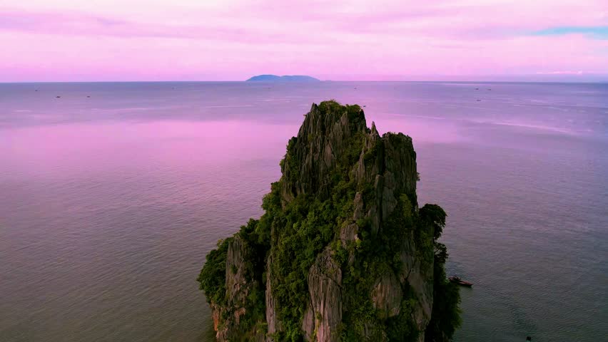 Drone shot of a lone limestone peak rising from the sea in Kien Giang, Vietnam, bathed in pink sunset light, creating a serene and tranquil seascape.