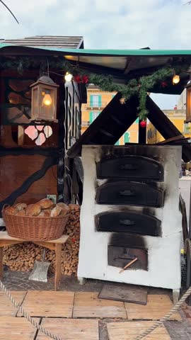 Rustic wooden Christmas market booth selling artisanal bread and pastries in woven baskets. Oven with stacked firewood, hanging lantern, and festive decorations create cozy holiday atmosphere