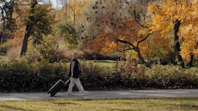 Senior woman with short hair walking on a path, pulling a wheeled suitcase in a beautiful autumn park - Powered by Shutterstock - Get 15% off with code: PIKWIZARD15